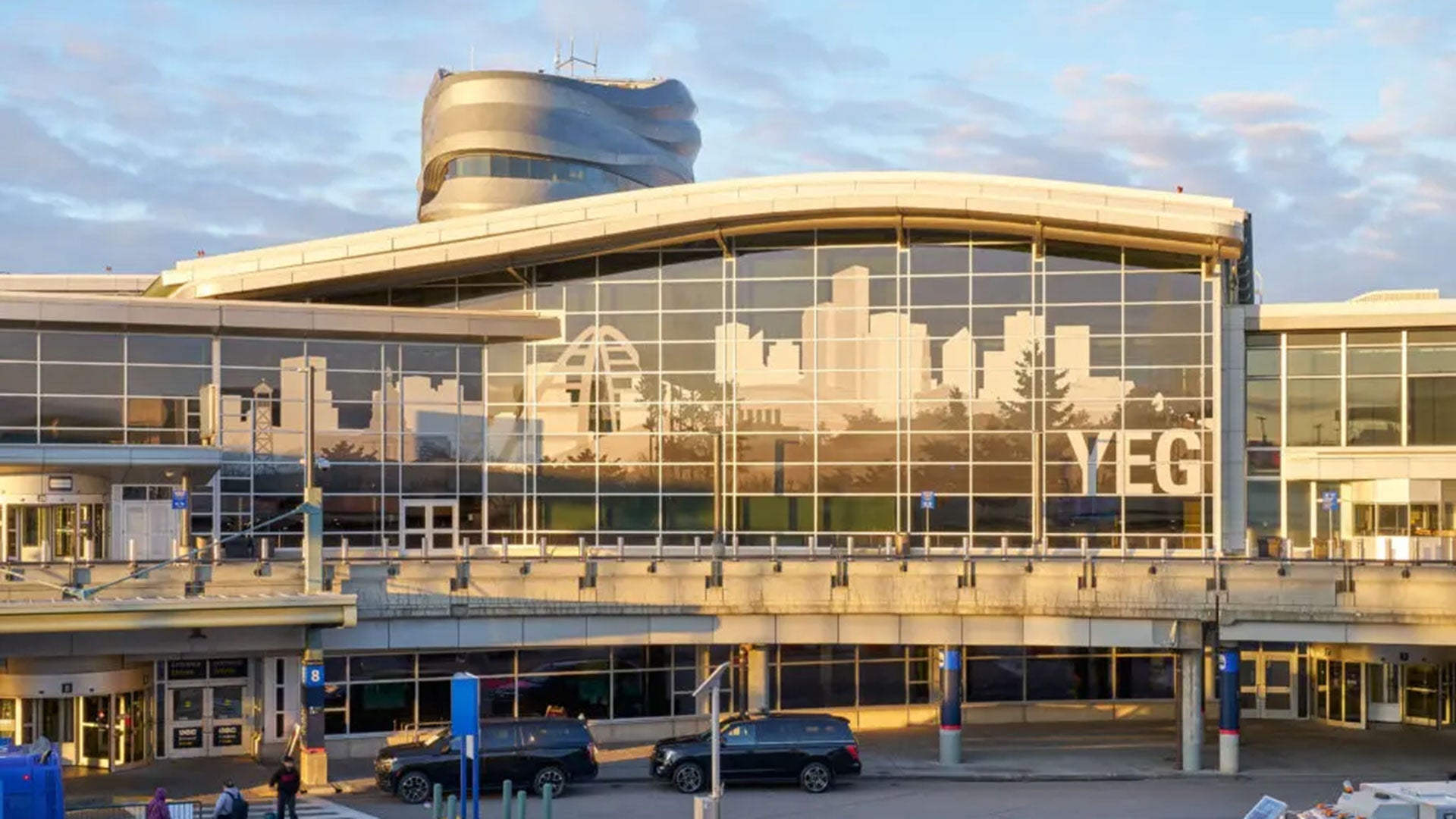 Exterior view of Edmonton International Airport departures terminal, featuring a large glass facade with a city skyline graphic and the letters “YEG” displayed at sunset.