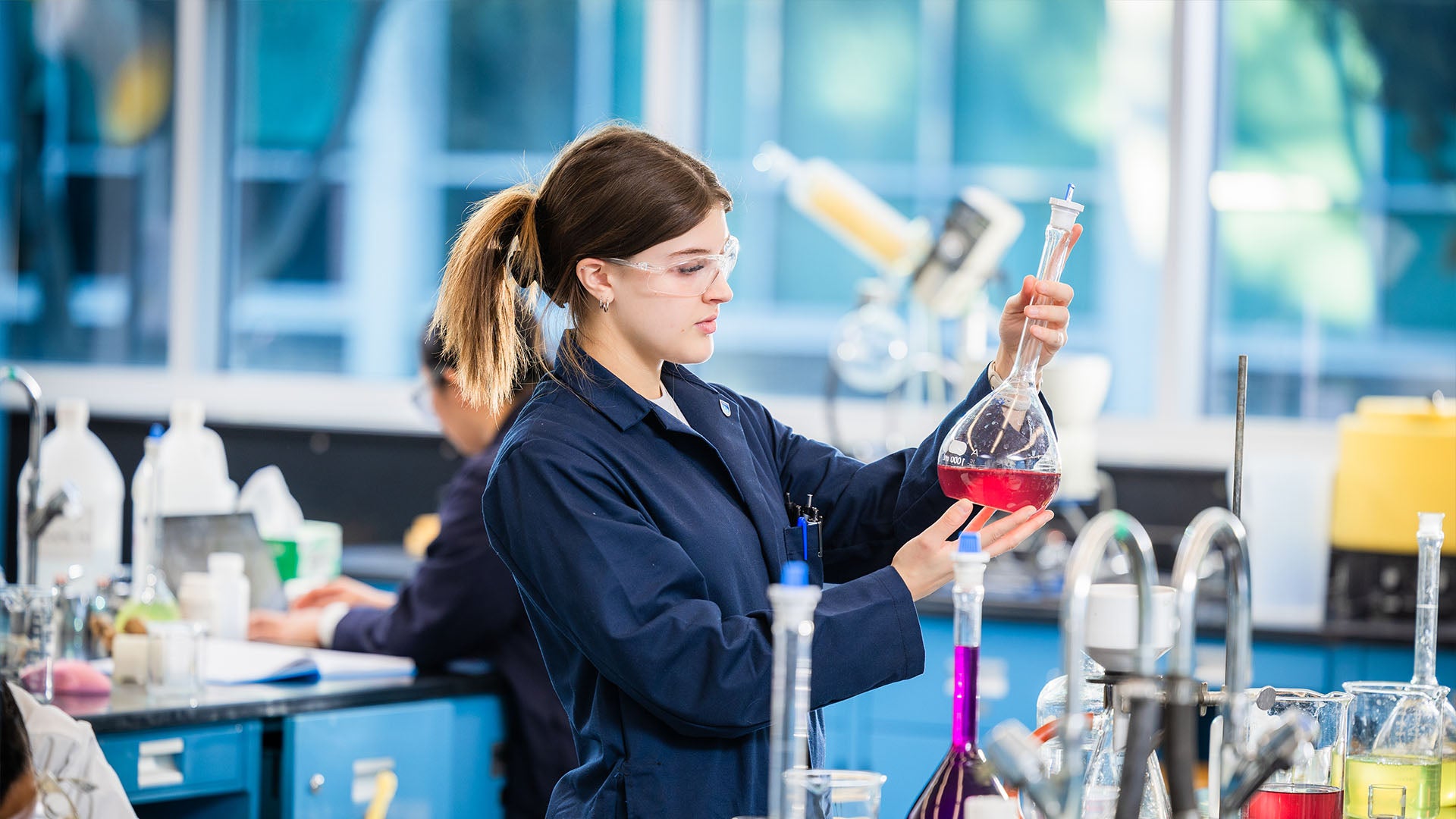 Chemical Technology student wearing safety glasses holds large flask containing a red liquid.