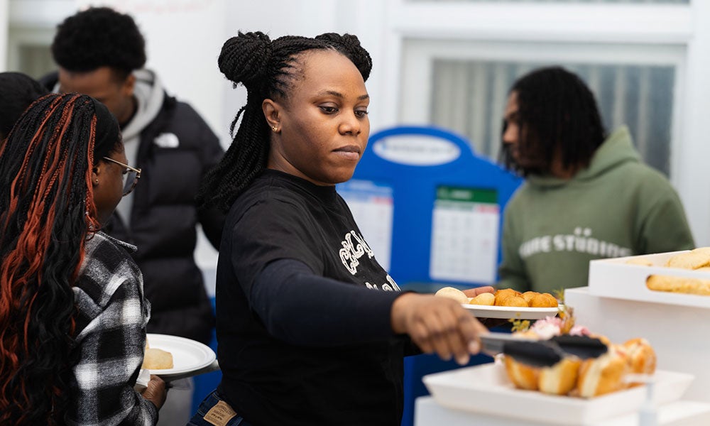 Person serving food with tongs from trays at a community gathering while several attendees stand nearby holding plates indoors.