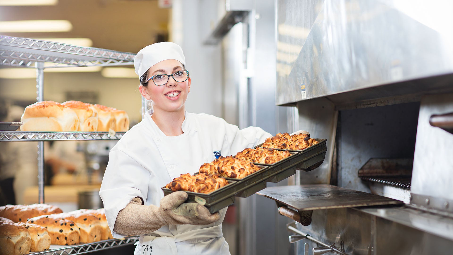 A smiling culinary student in a professional kitchen holding a tray of freshly baked bread, wearing an apron, chef’s uniform, and oven mitts.