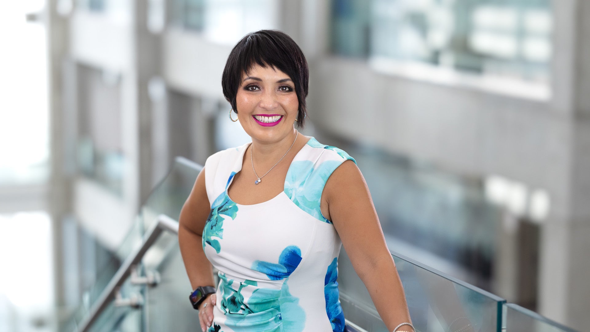 Waist-up portrait of a person with short dark hair in a white floral dress, standing by a glass railing in a bright modern office with a softly blurred background.