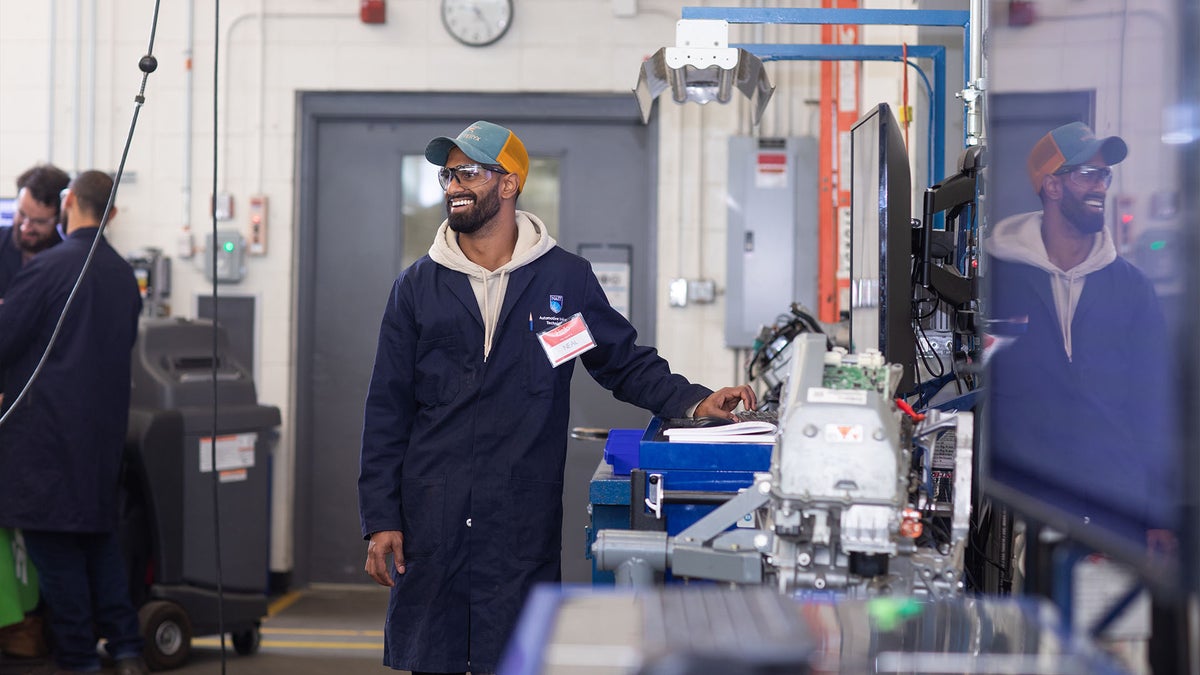 A man wearing a baseball cap, blue work coat, and safety glasses smiles while standing in an auto repair shop.