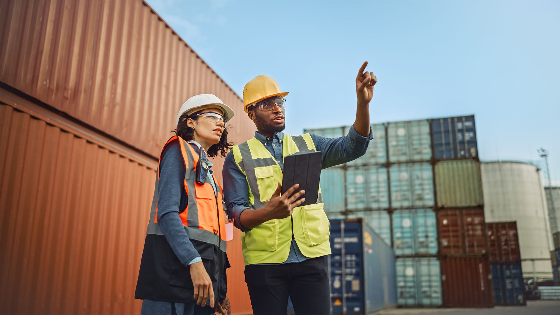 Two logistics professionals in safety gear, including hard hats and reflective vests, discussing operations in a shipping container yard.