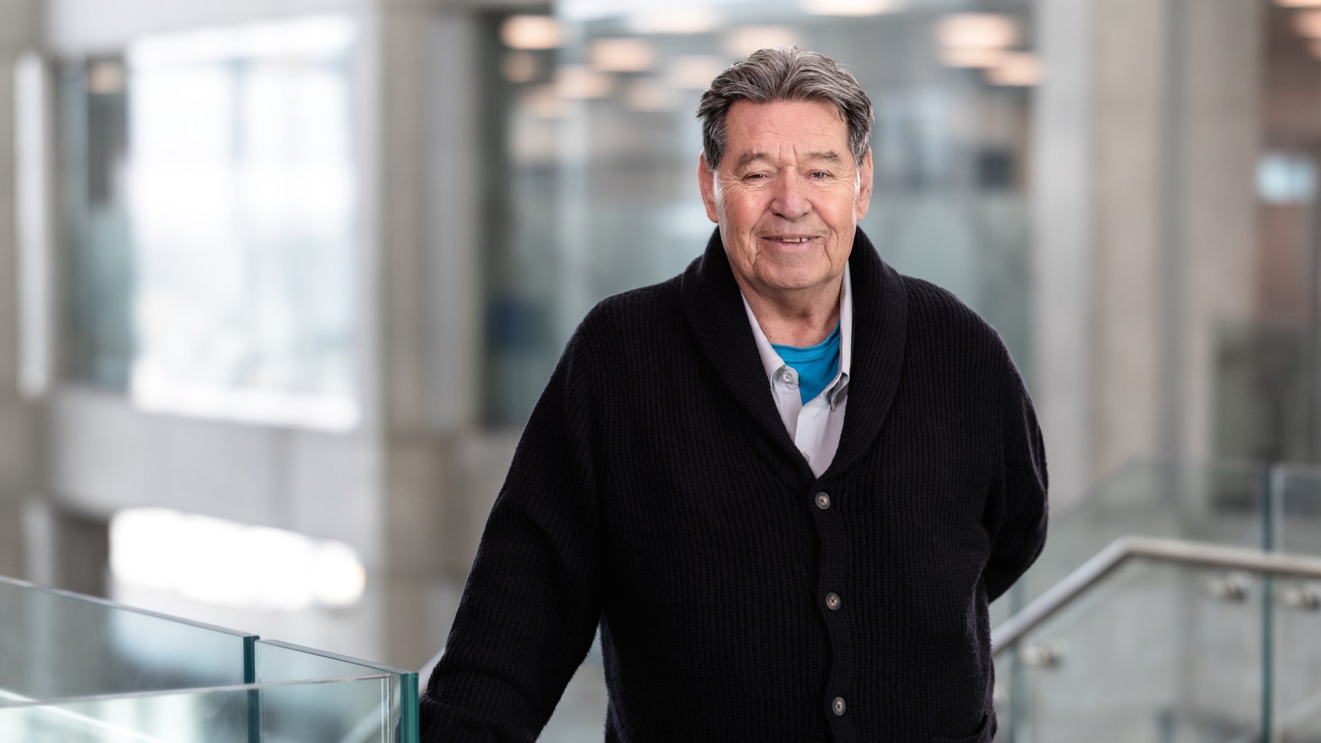 Head-and-shoulders portrait of a person with short gray hair wearing a dark sweater, positioned indoors in a bright modern space with a softly blurred background.