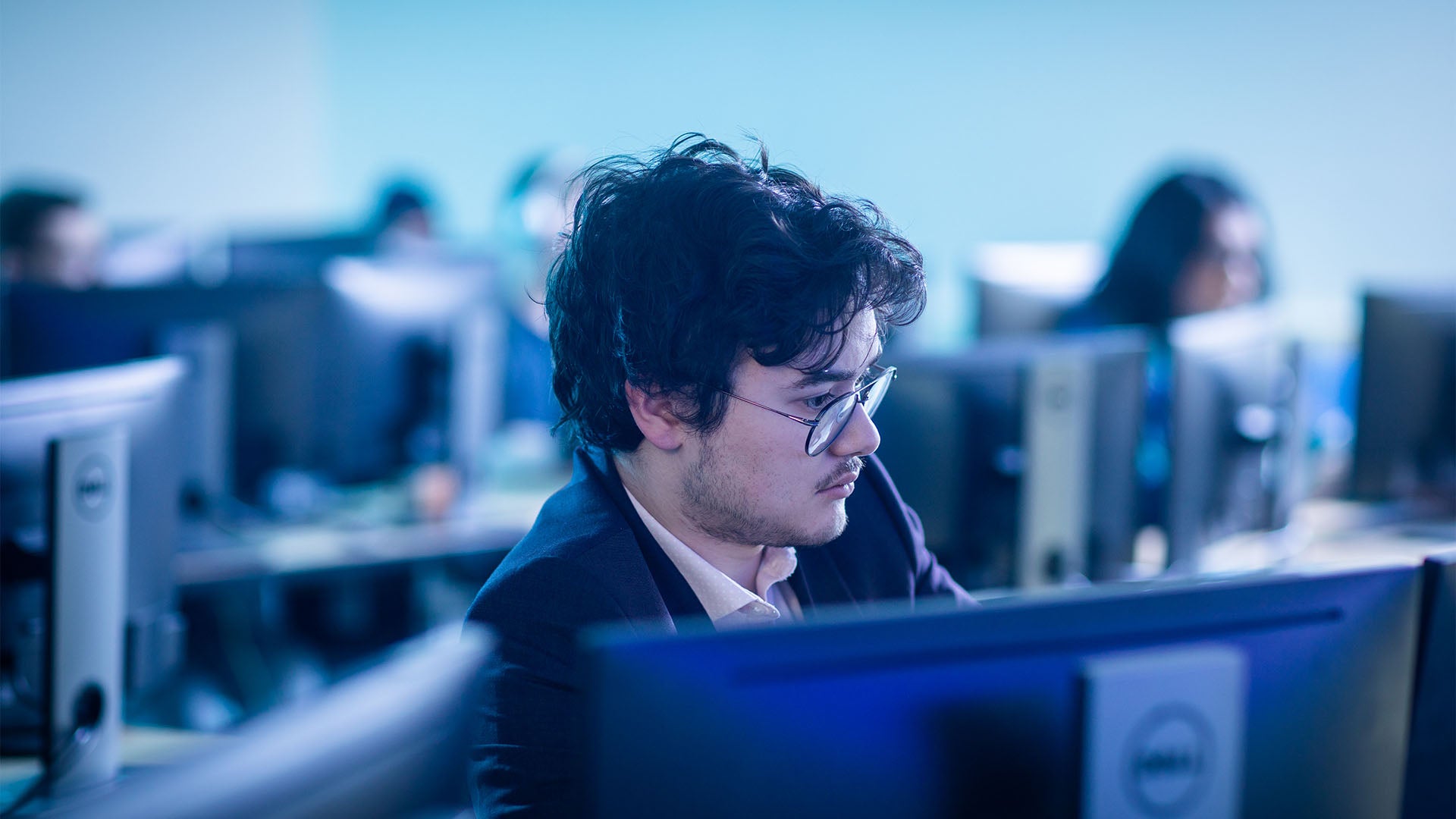 Man with glasses concentrating on a computer in a computer lab surrounded by other students at separate stations.