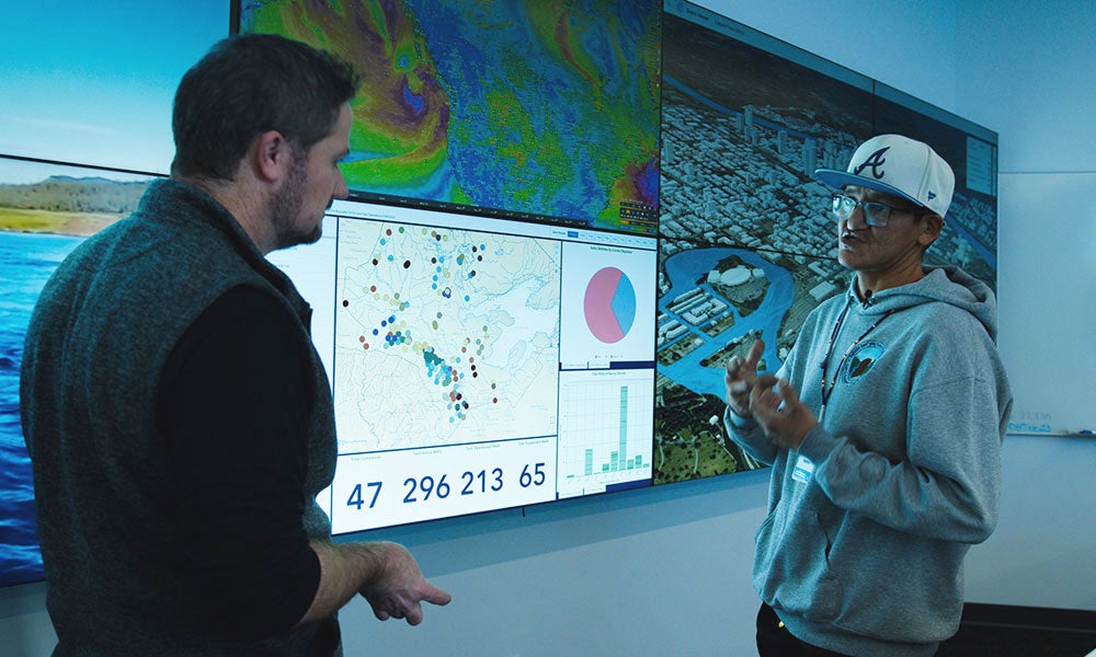 A man in a hat talking to another man in a room with a large screen