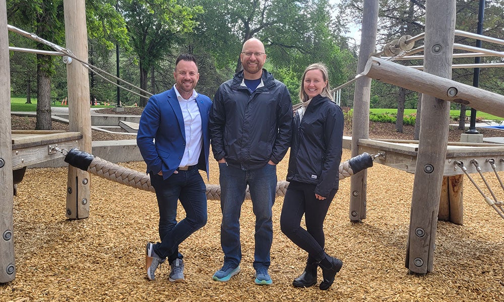 Three people stand on a wood-chip playground surface among large rope-climbing structures and timber play equipment in a park setting.