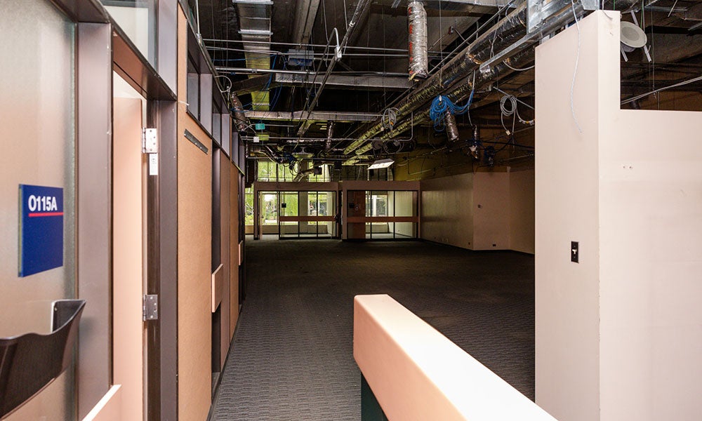 Interior space under renovation with exposed mechanical and electrical systems, enclosed rooms along one side, and an open area leading to glass exit doors.