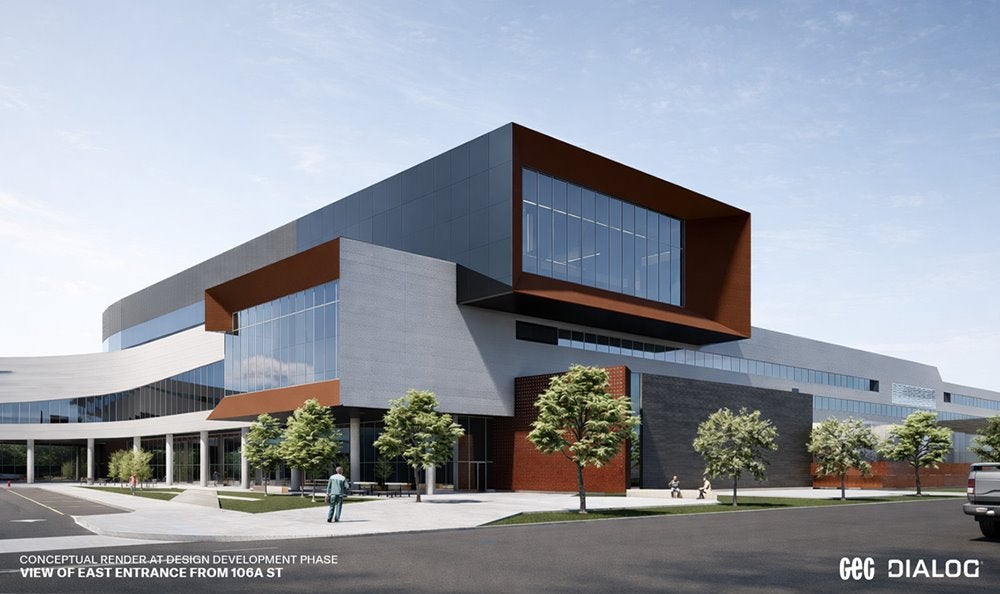 Modern, multi-storey institutional building with a mix of glass, white, and red-brown exterior panels, featuring large angular overhangs and expansive windows. The view shows the east entrance from a street corner, with a sidewalk, landscaped trees, and a few pedestrians nearby. 