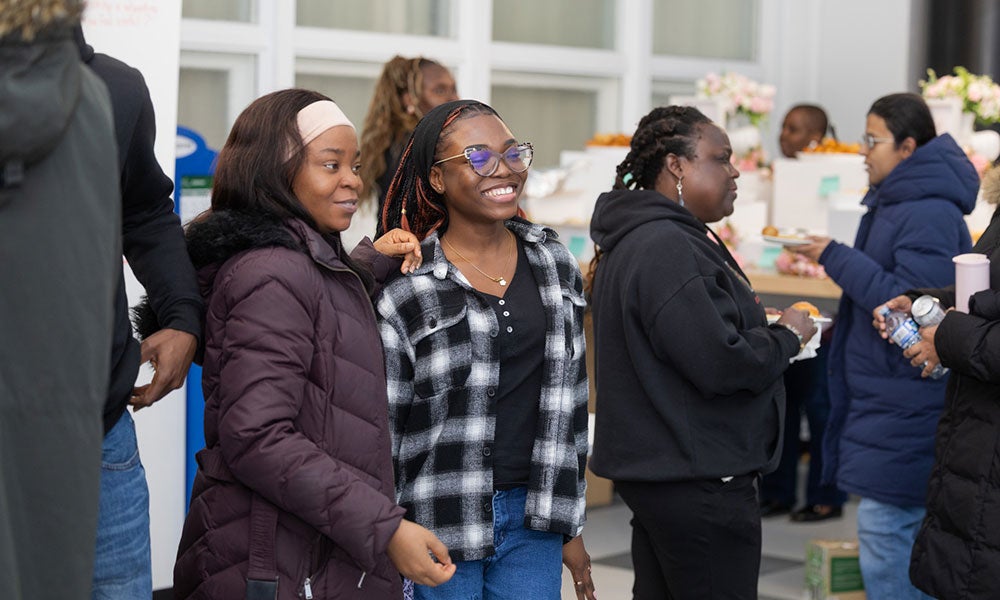 Several people gather and converse at an indoor community event, with attendees standing near food and refreshment tables in the background.