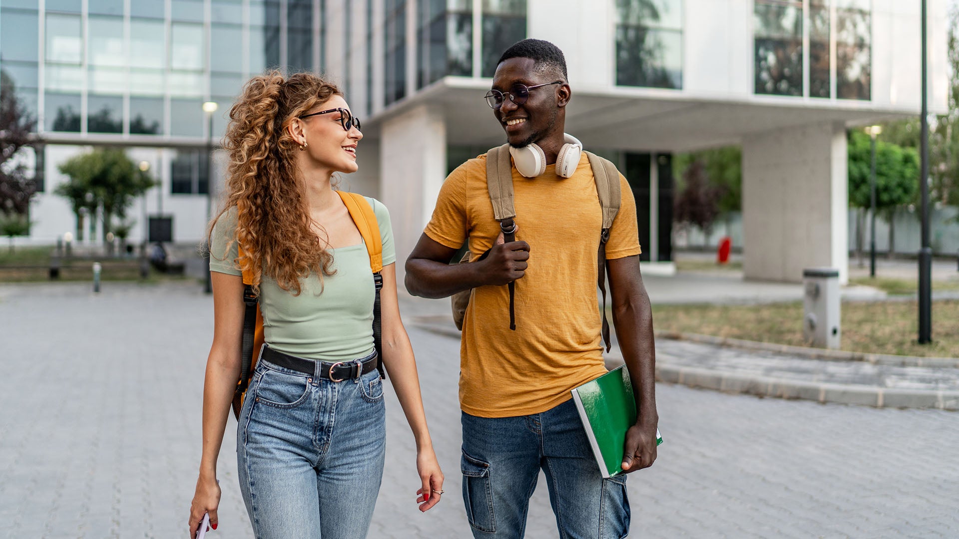 Two students wearing back packs, smiling at each other as they walk outside a school.