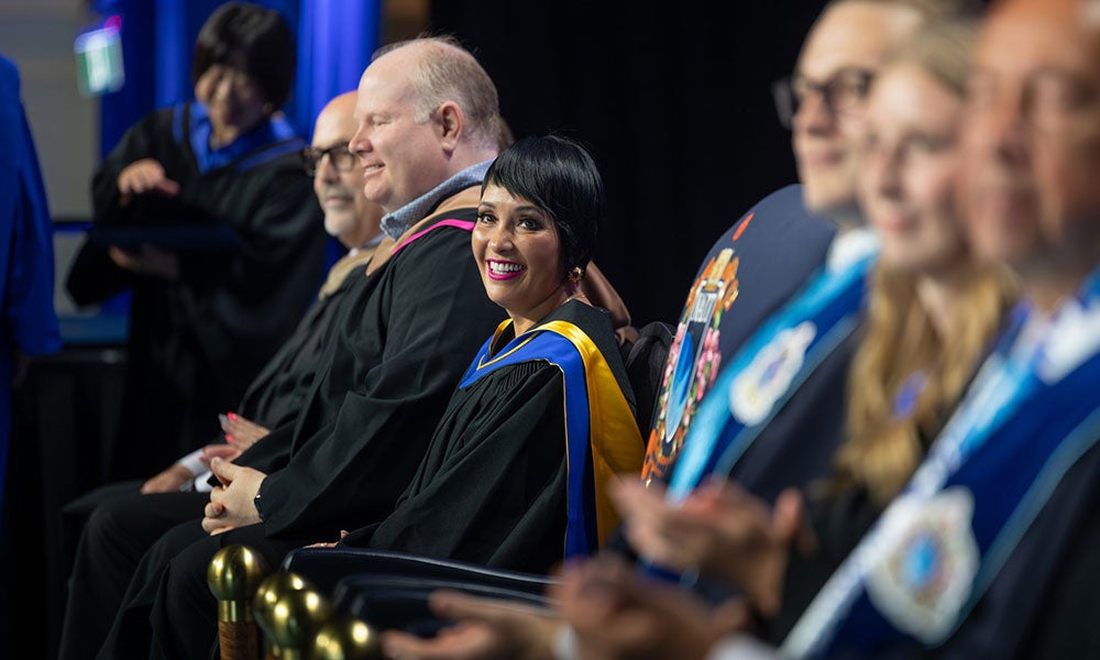 Seated faculty in academic robes on stage, with one person in the center turning toward the camera and smiling during a graduation ceremony.