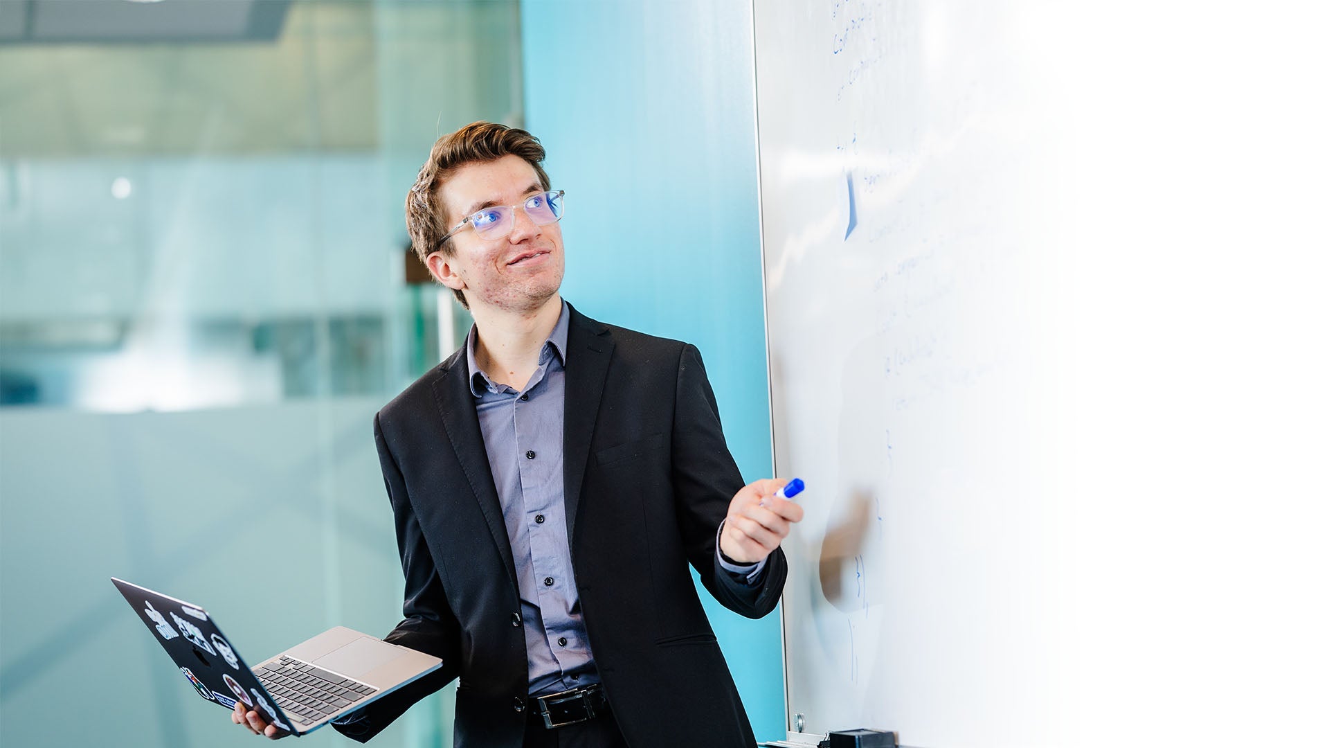 A man wearing glasses and a suit, holding a laptop with one hand and a marker in the other, pointing at and looking at a whiteboard.