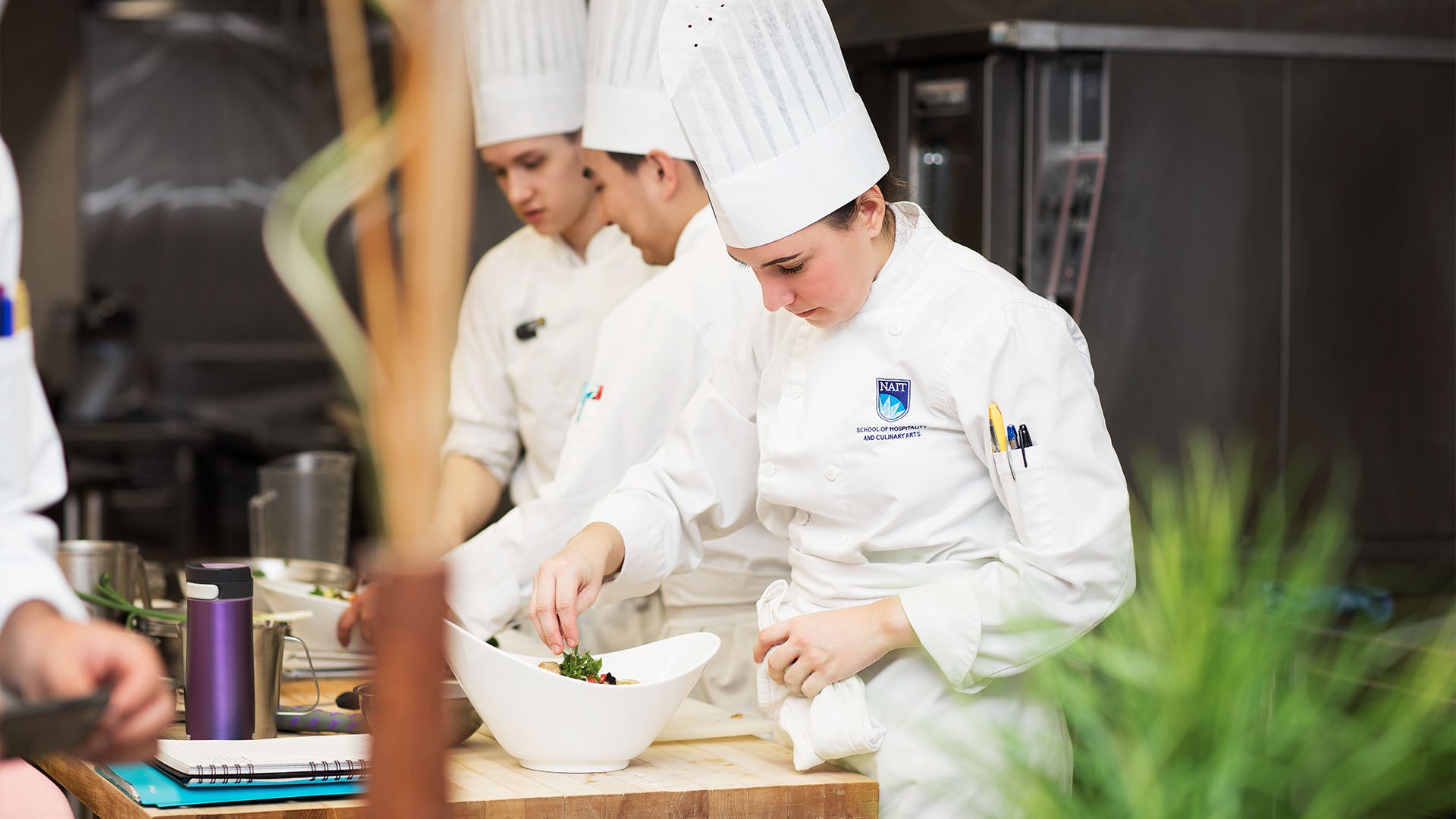 A culinary student in a professional chef uniform and toque garnishing a dish, with two other students focused on their tasks in the background.
