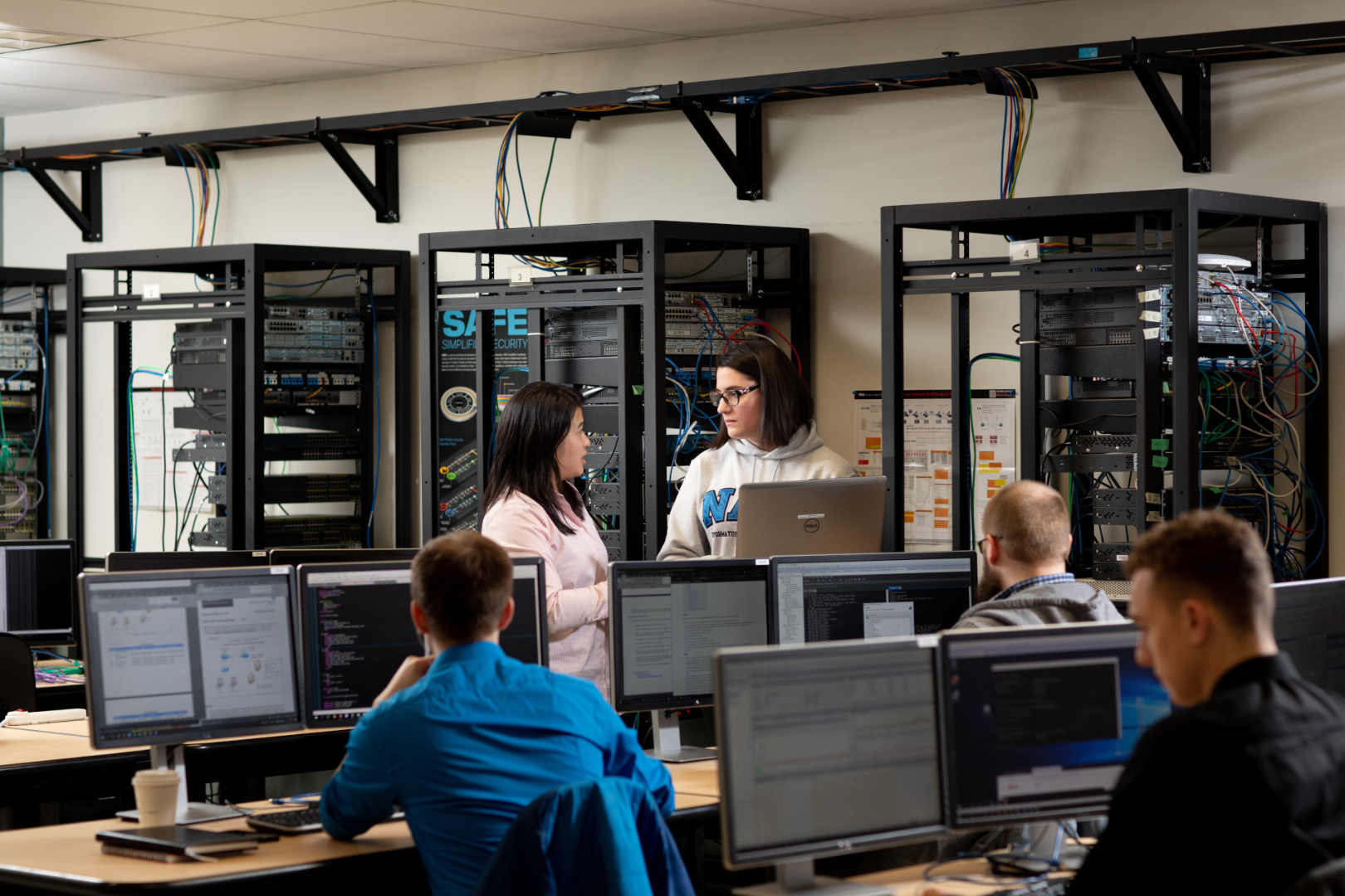 Students work at computer stations in a network lab equipped with server racks and exposed cabling. In the centre of the room, two people discuss work while looking at a laptop, with multiple monitors displaying code and technical applications in the foreground.