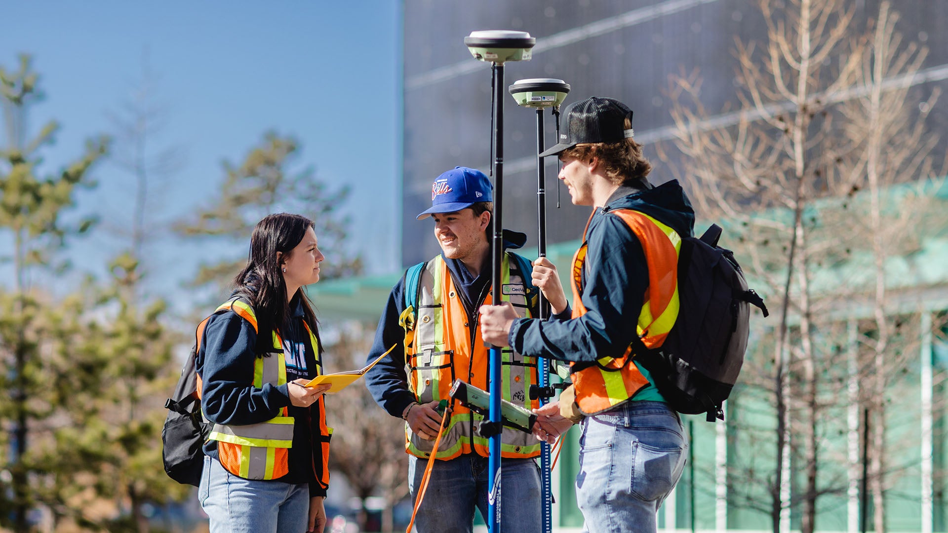 Three surveying and geospatial engineering technologists wearing high visibility safety vests and holding equipment to map and measure surrounding features.