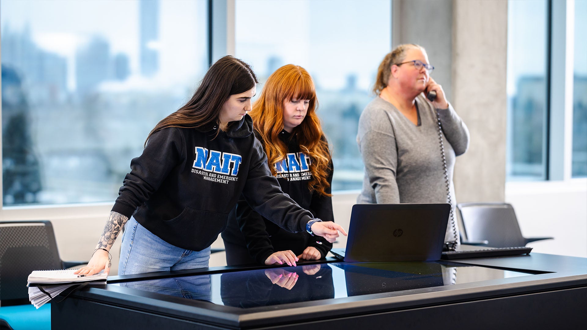 Two students wearing NAIT branded hoodies use a laptop on top of an interactive tabletop while a woman in the background speaks on a phone.