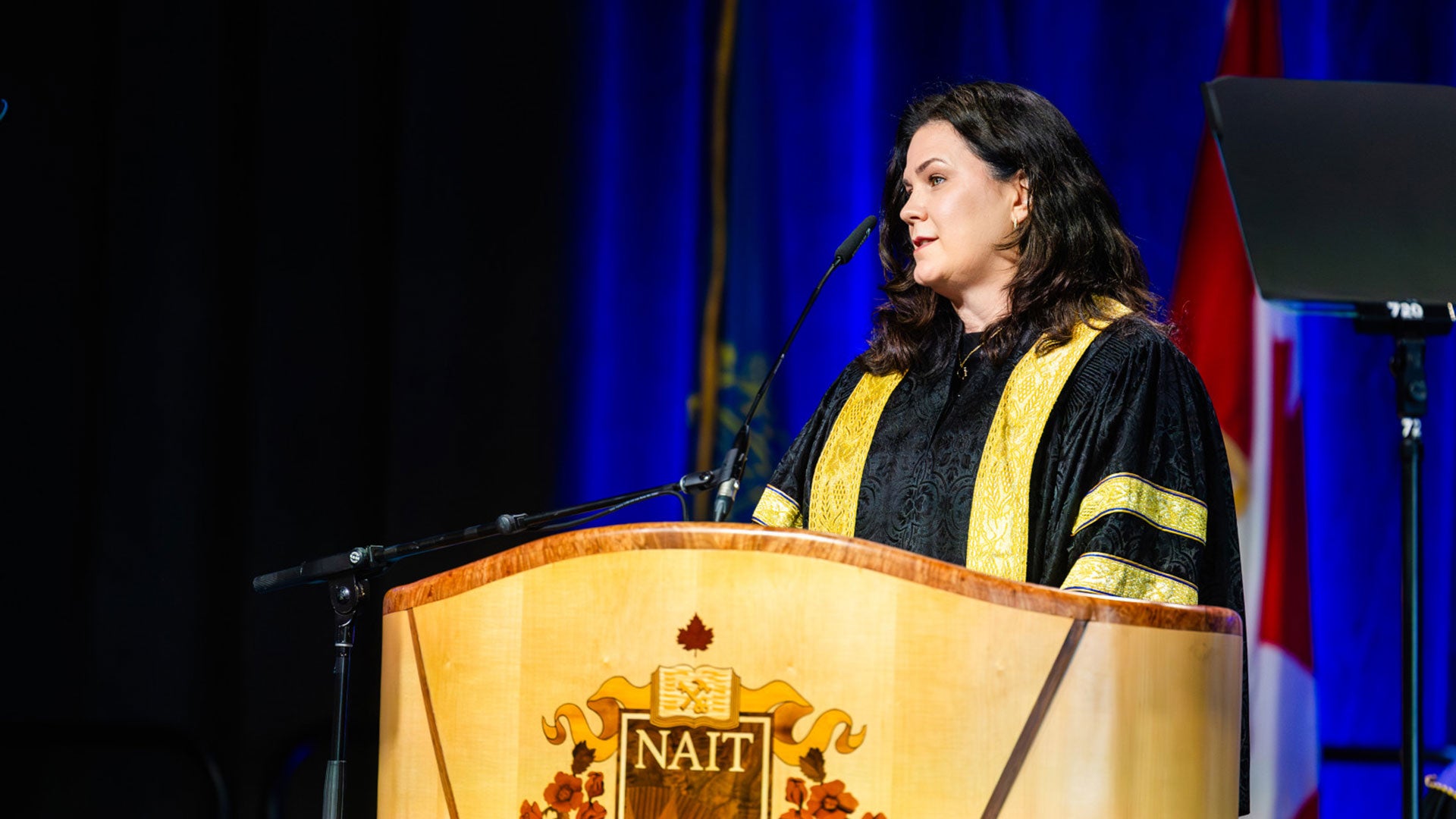 Person speaking at a NAIT convocation podium on a stage with blue curtains and a Canadian flag in the background.