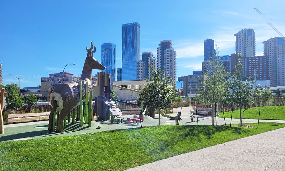 Deer-themed playground with climbing structures and slides in a landscaped urban park, set against a backdrop of downtown high-rise buildings and a clear blue sky.
