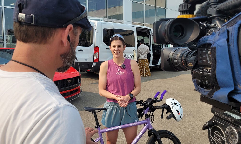 Person standing beside a purple bicycle while being filmed by a camera crew outside a building, with vehicles visible in the background.