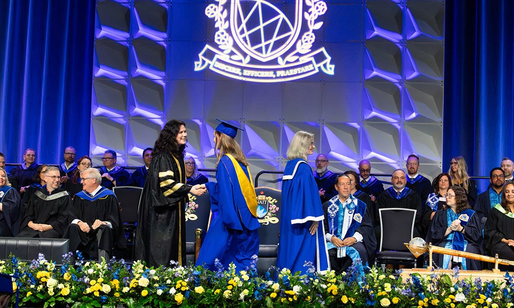 Graduates cross the stage during a NAIT convocation ceremony while faculty and officials are seated in the background.