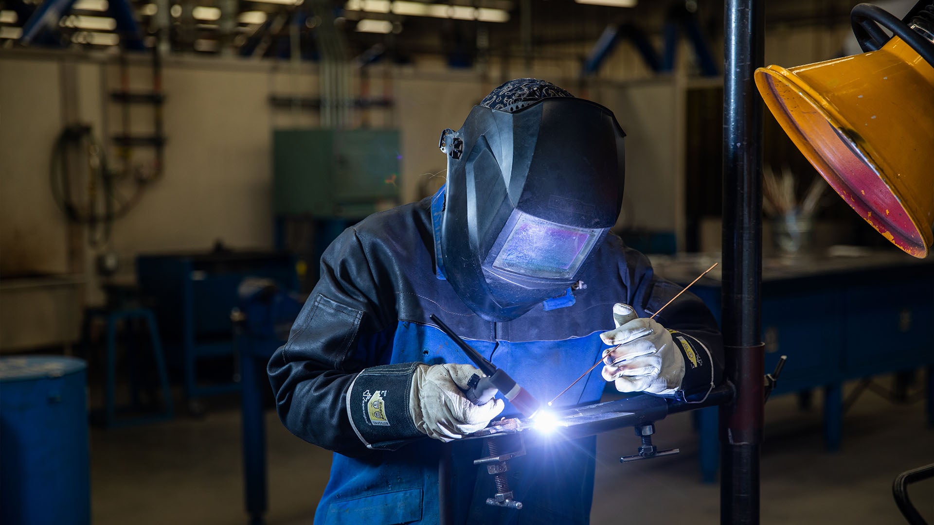 A welding student wearing a protective welding mask and jacket while performing a weld in a workshop.