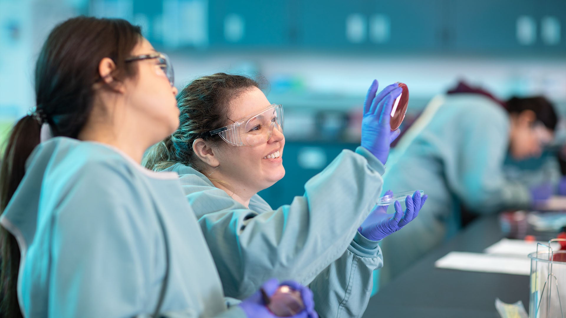 Two female students wearing gowns and safety glasses view a petri dish.