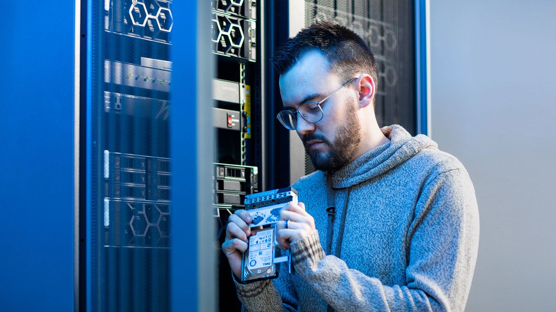 Man in a server room working on hardware, holding a hard drive near a rack of servers, wearing glasses and a sweater.