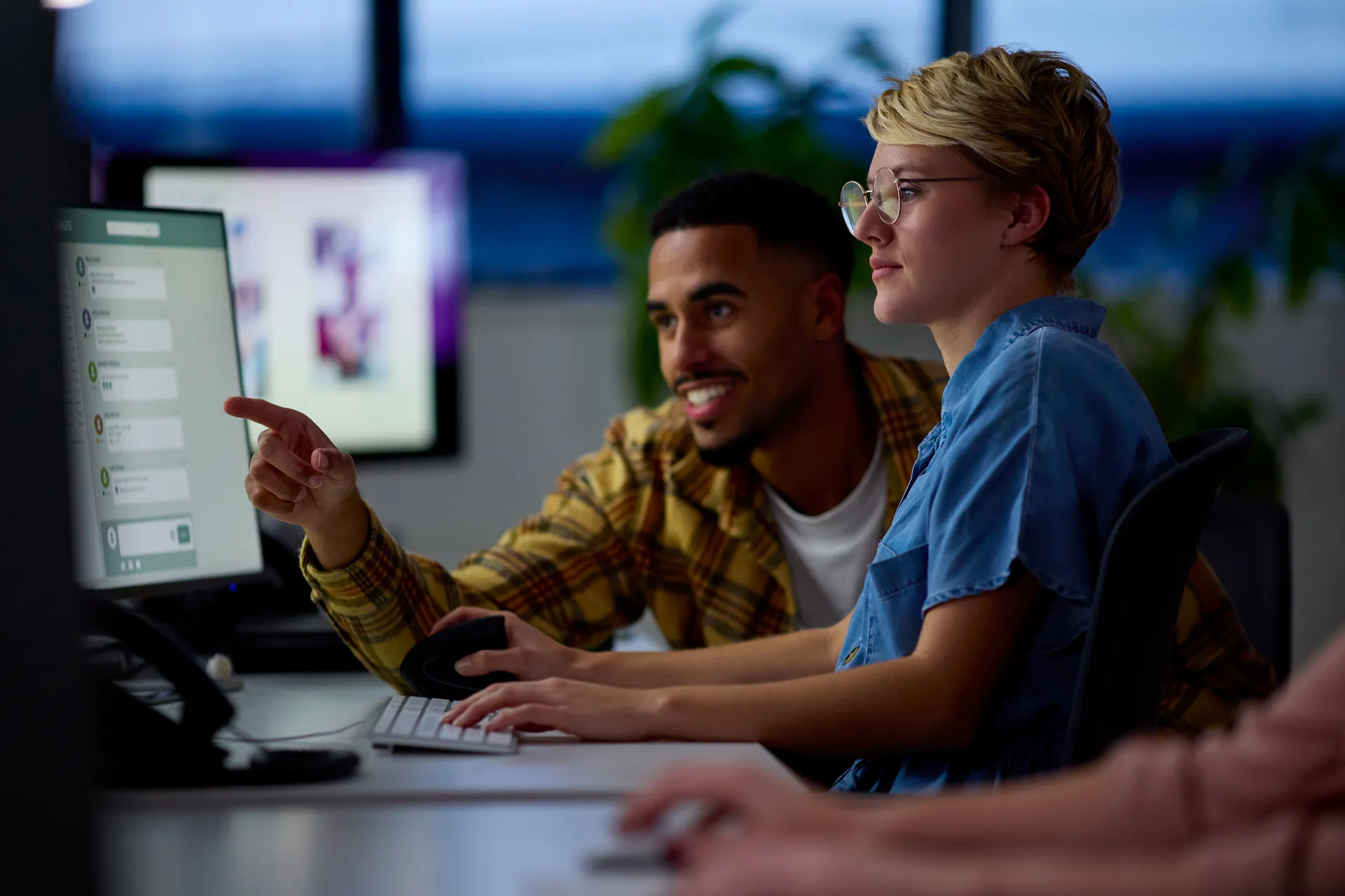 Two Business Colleagues At Desks In Office Working Late On Project Together