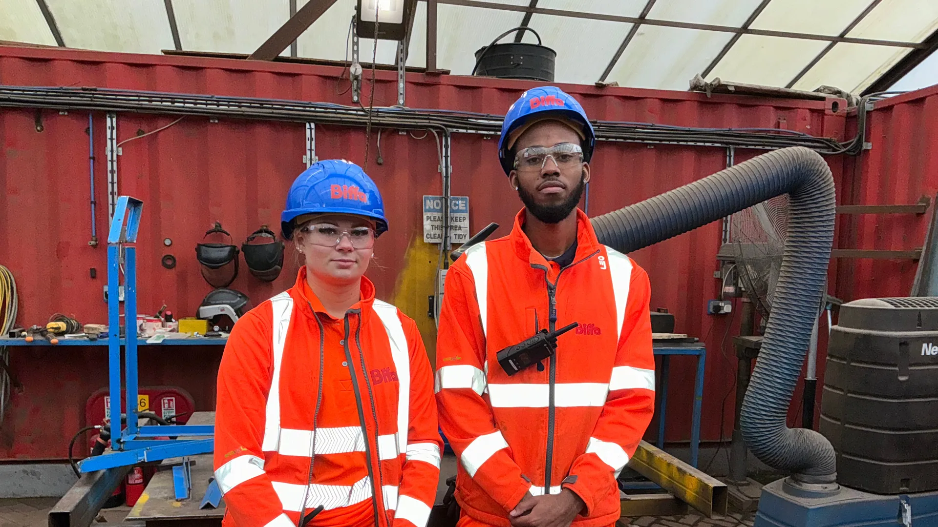 Two young people on a work site wearing orange high-vis outfits and blue hard hats which have the Biffa logo