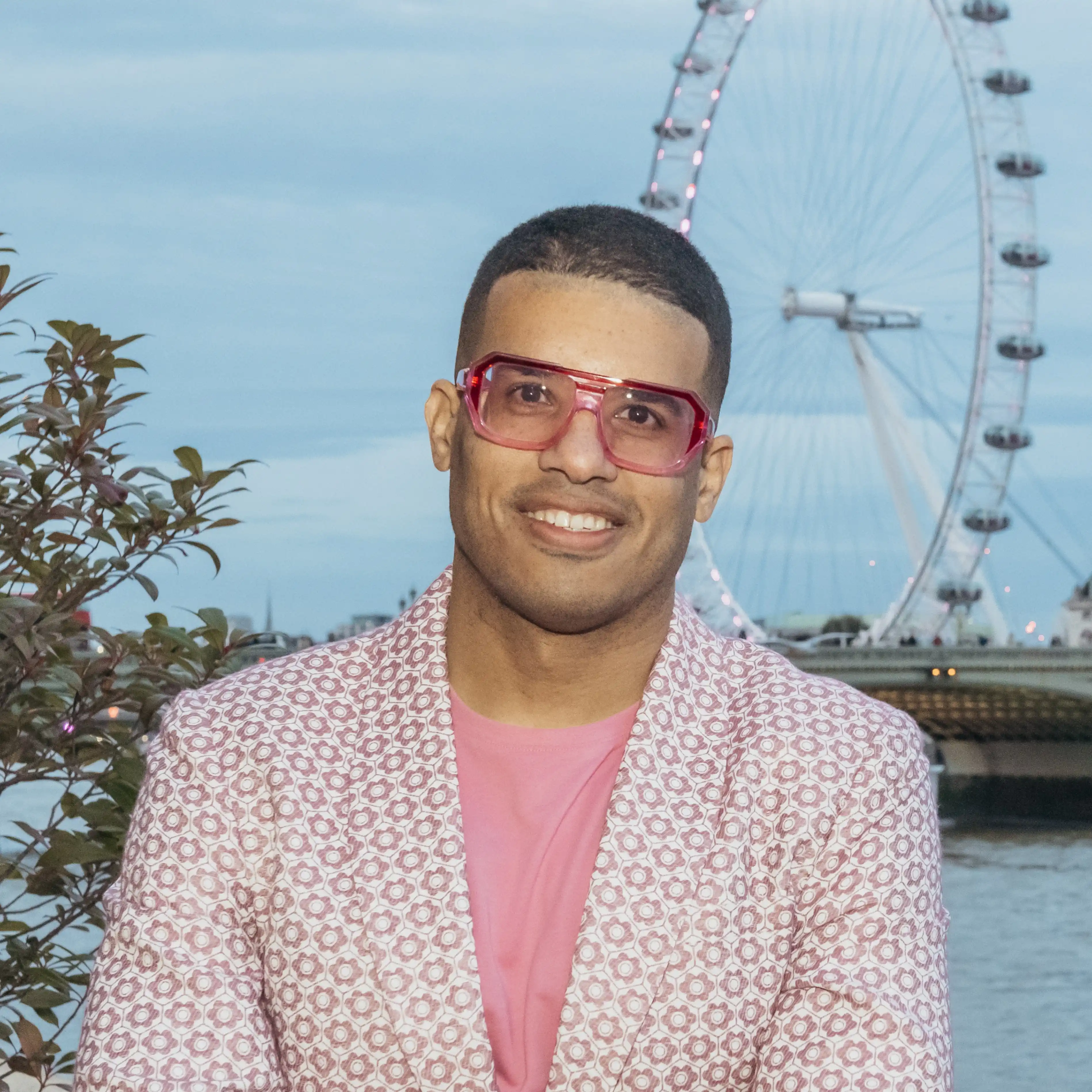 A young male wearing a pink and white jacket and pink glasses, with a ferris wheel in the background