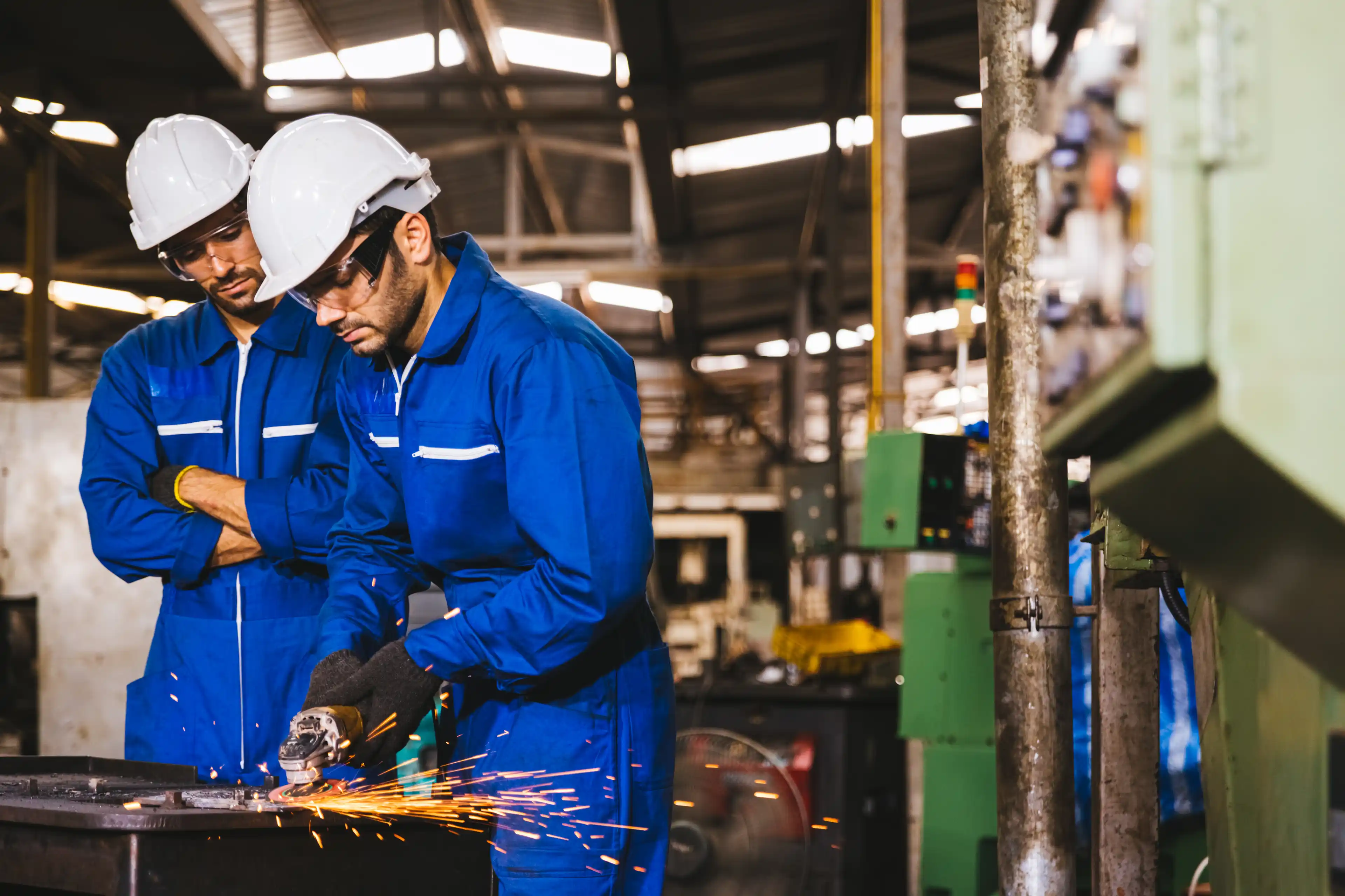 Group of technician industrial engineers wearing safety uniform and safety helmet cutting metal part using hand angle grinder machine. Large industrial factory background.