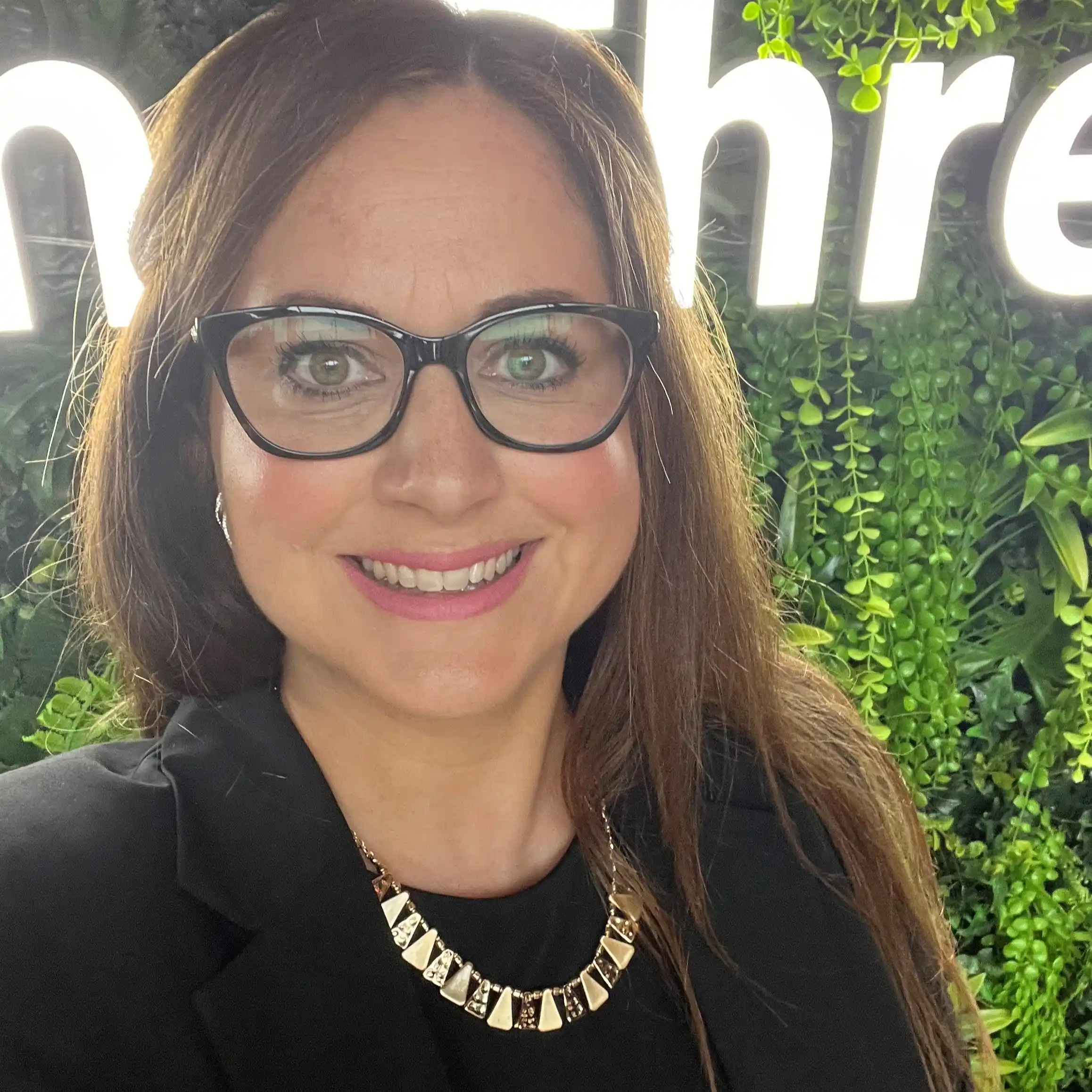 A dark haired female wearing black top with necklace and black glasses
