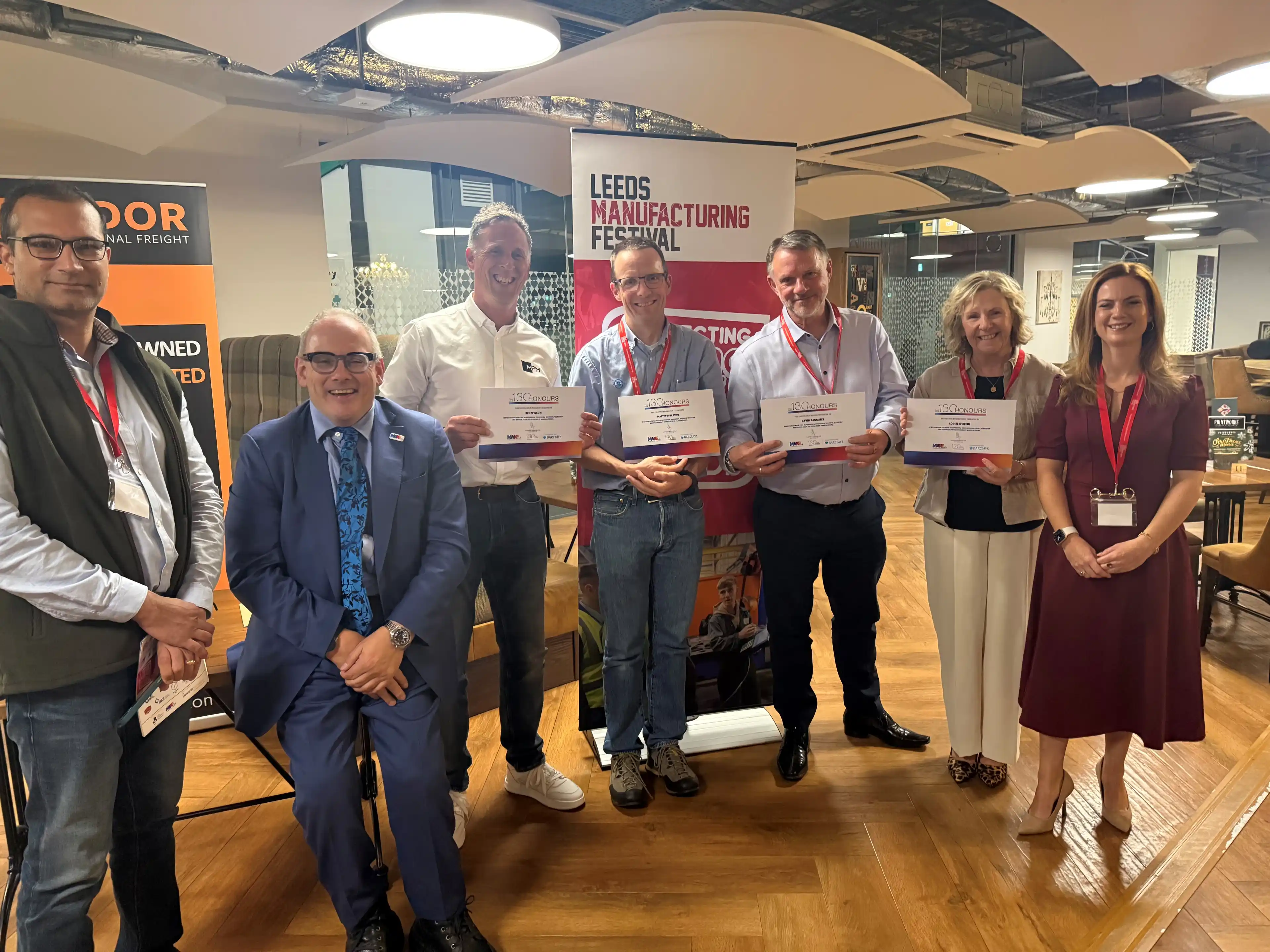 7 people holding certificates posed in front of roller banners displaying Leeds Manufacturing Festival