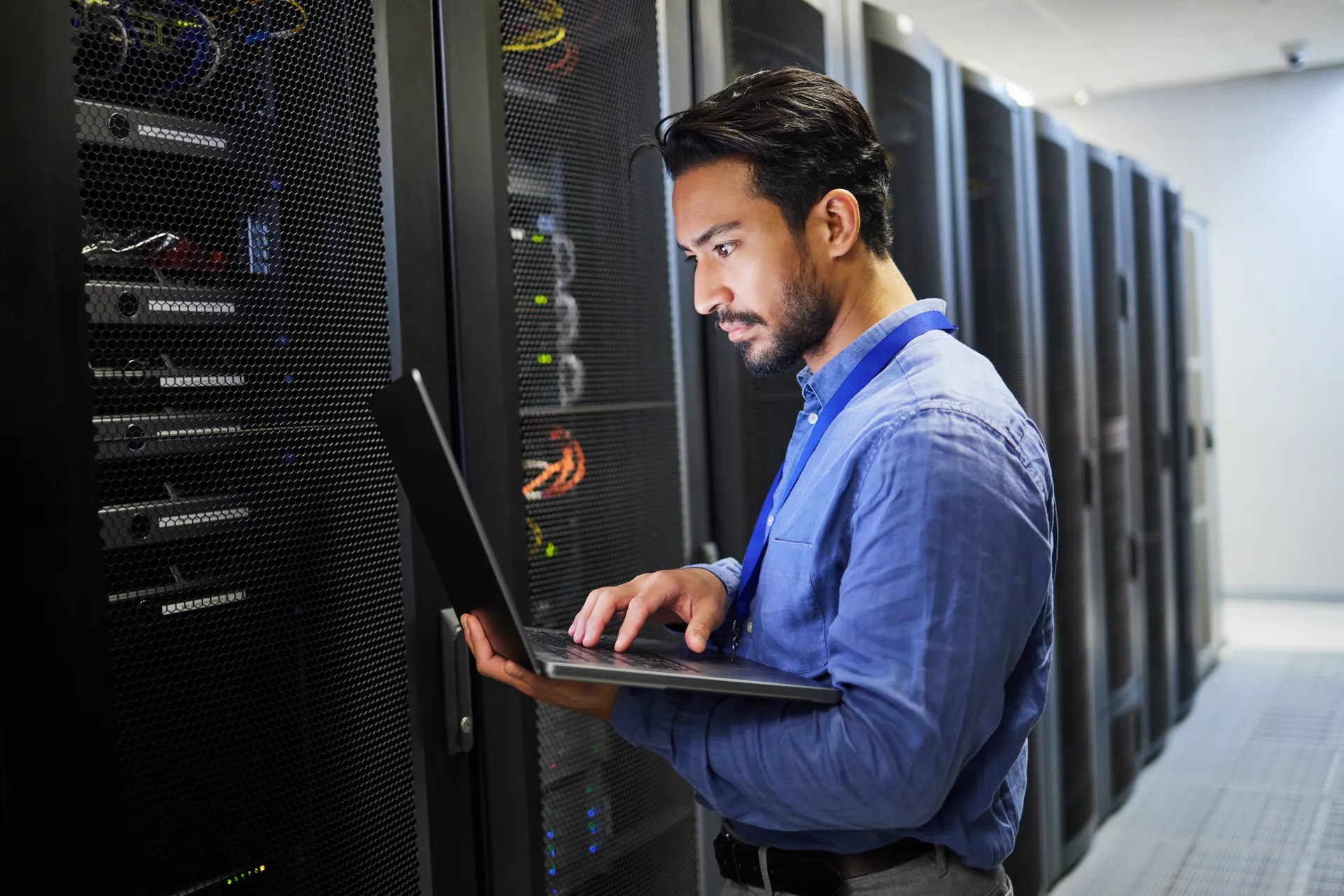 man with blue shirt in server room looking at laptop screen