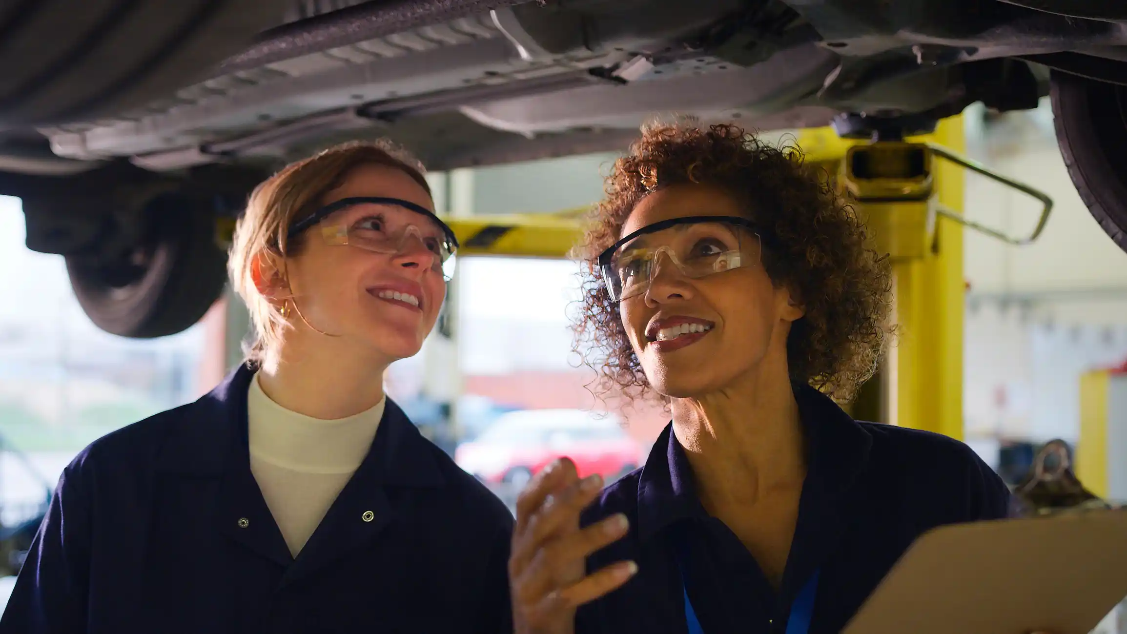 Female Tutor With Student Looking Underneath Car On Hydraulic Ramp On Auto Mechanic Course