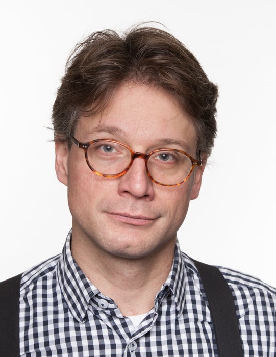 Portretfoto van een man met bruin haar en een lichtbruine bril gekleed in een overhemd met zwart-witte ruitjes en zwarte bretels