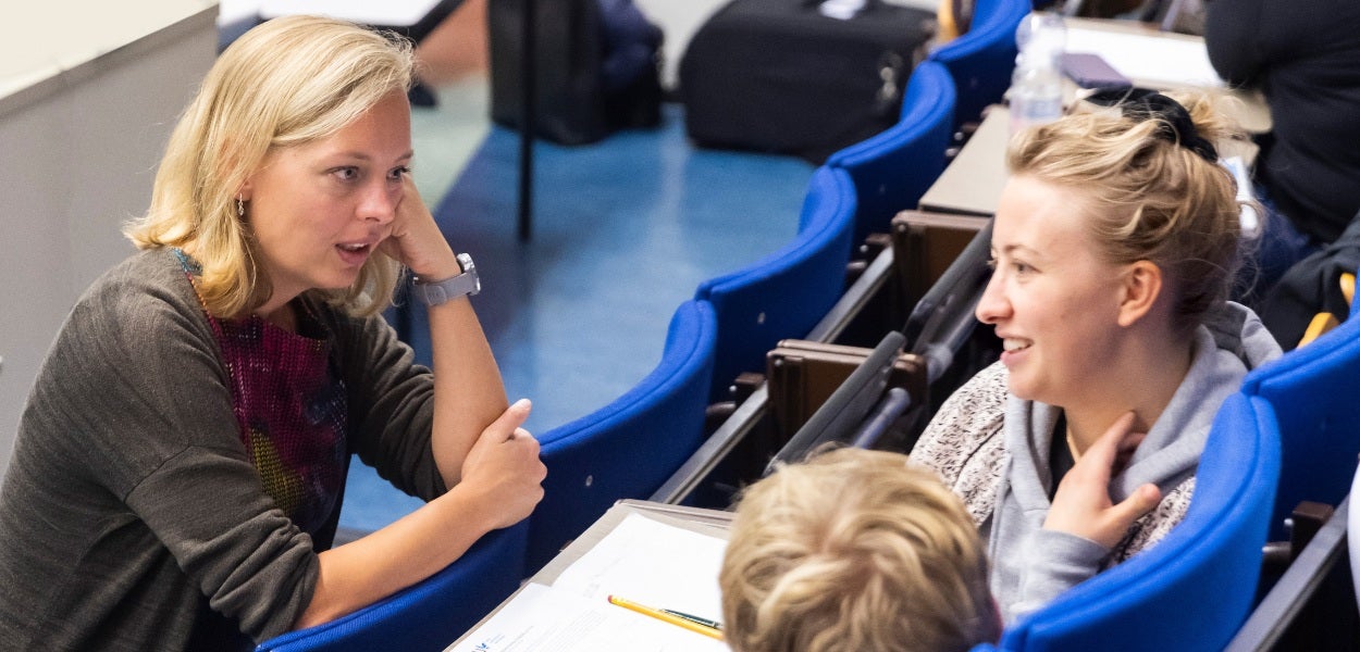 Two blonde women sit across from each other in a lecture hall with blue chairs.