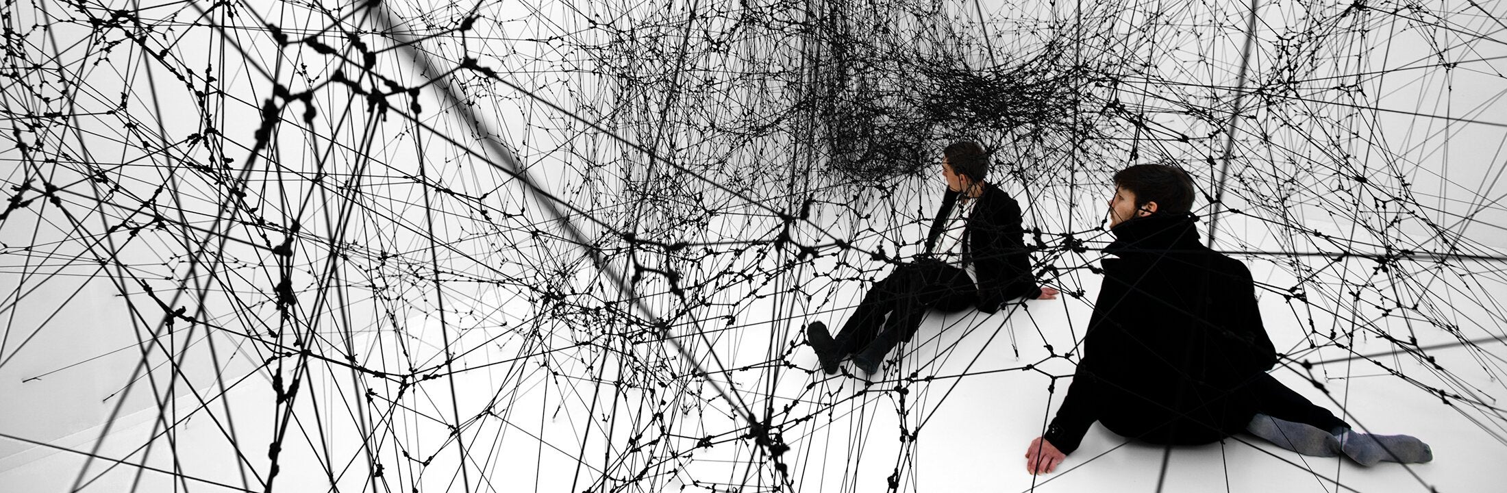 Twee mannen zitten in een gigantisch, zwart spinnenweb (sculptuur van Tomas Saraceno) 