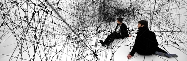 Twee mannen zitten in een gigantisch, zwart spinnenweb (sculptuur van Tomas Saraceno) 