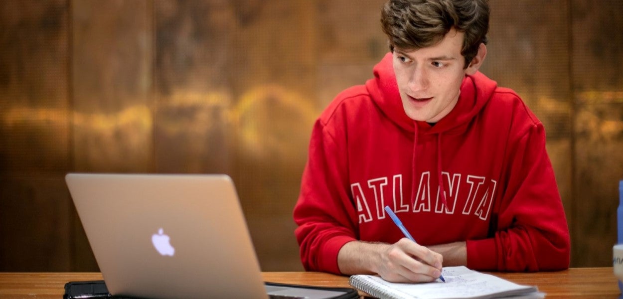 A student in a red sweat shirt looks at a laptop and writes on a notepad