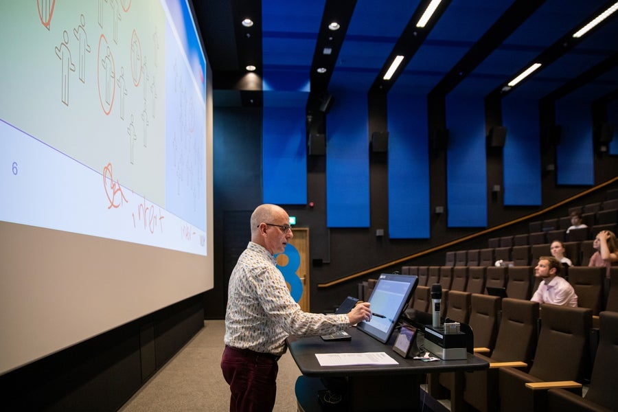 Teacher in a lecture hall in front of a large screen showing a presentation