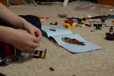 Small hands working with lego on the floor, the explanation in front of them, surrounded by more lego. 