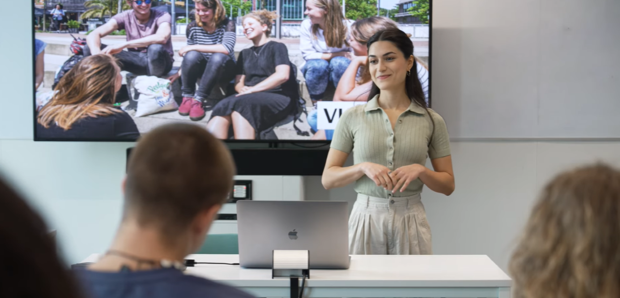 Een docent staat voor een beeldscherm en kijkt glimlachend de studenten in de zaal aan