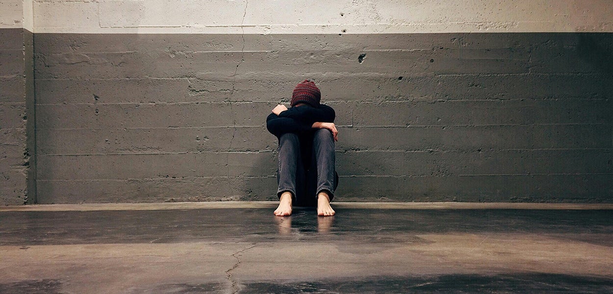 A young person sits looking dejected and hunched over in a gloomy room.