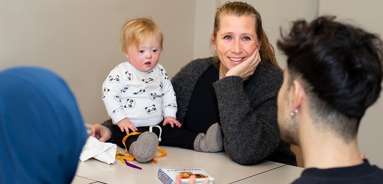 A woman offers a listening ear to parents, she has her arm around a young child sitting on the table. Photo: Erik Buis