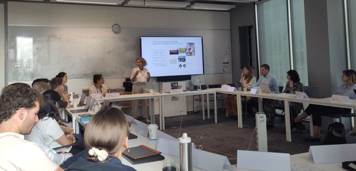 The image shows a group of people sitting around a long table in a conference room. A woman is standing at the head of the table giving a presentation, and a projector screen displays her presentation slides. The room appears to be a modern, well-lit office space, suggesting a professional setting, possibly a workshop or seminar. The participants are attentively listening to the presenter.