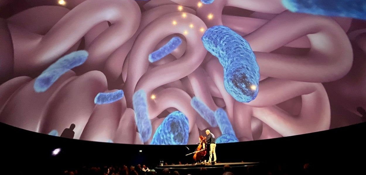 Een man spreekt op een podium en een vrouw speelt cello.