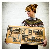 A woman with red-blond hair in a bun and glasses, dressed in a knitted jumper holds a wooden scale model of the VU campus in her arms