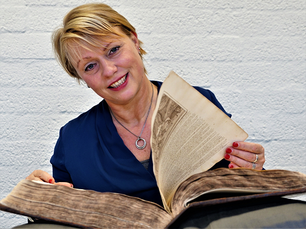 A smiling woman with short blond hair, wearing a dark blue blouse and red lacquered nails is leafing through an old Bible.
